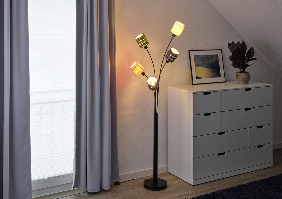 Lampadaire multicolore allumé contre un mur, à côté d'une commode grise et d'une fenêtre.