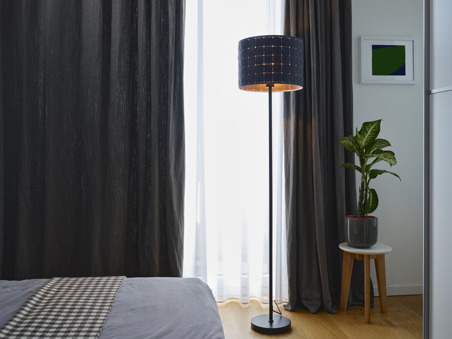 Lampadaire allumé dans une chambre, près de rideaux gris, d'une plante et d'un lit.