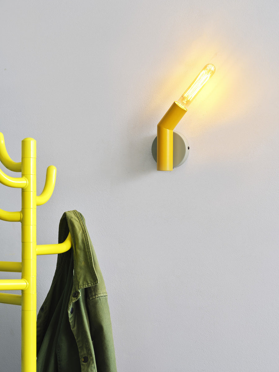 Gros plan de l’applique allumée sur mur blanc, ampoule filament et portemanteau flou.