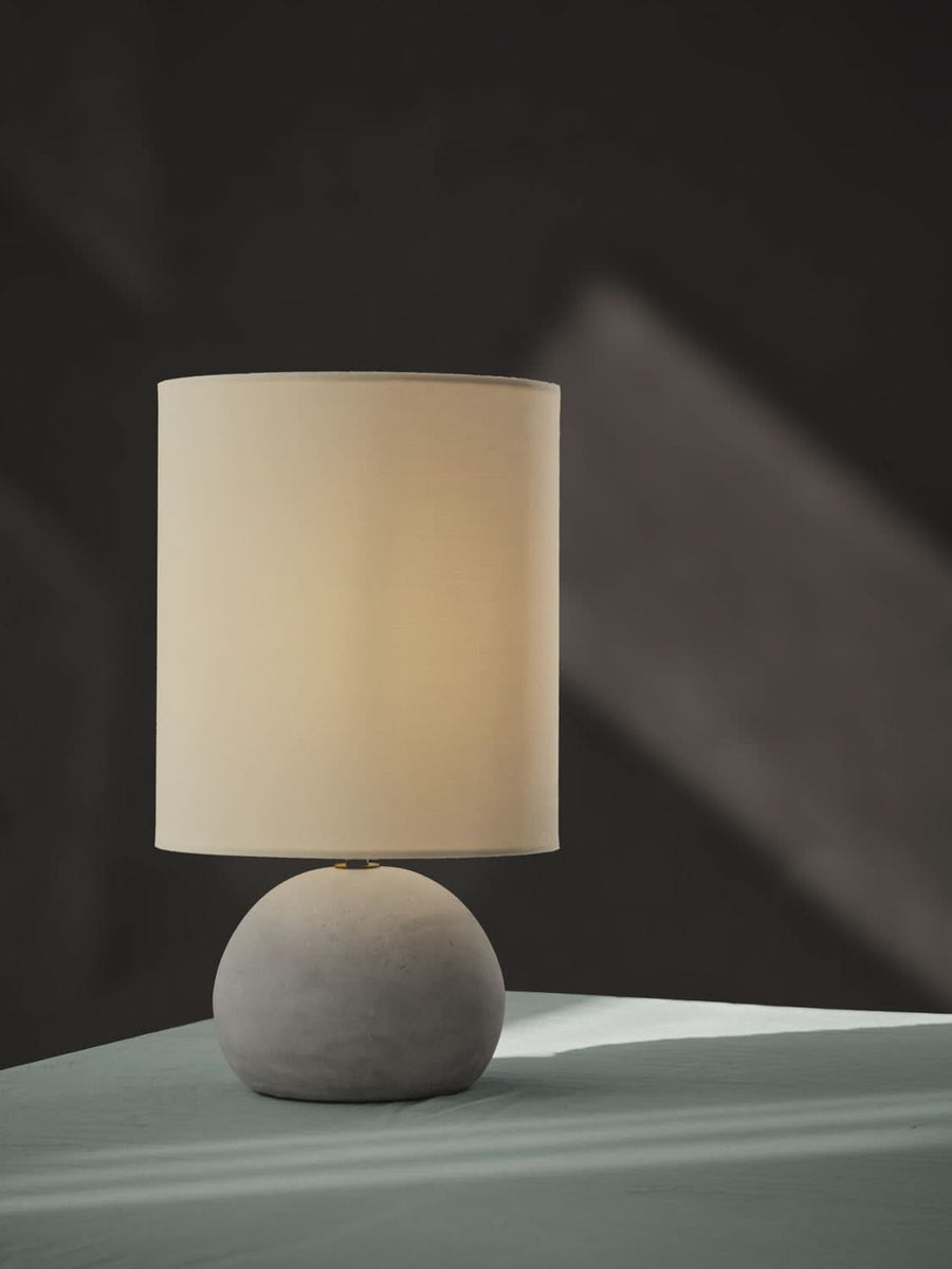 Vue de profil sur fond sombre, lampe allumée avec base sphérique en béton gris.
