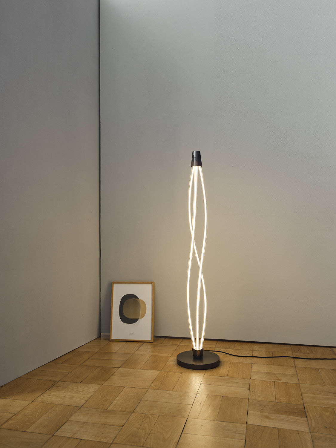 Lampadaire allumé dans un coin, près d'une affiche encadrée sur parquet.