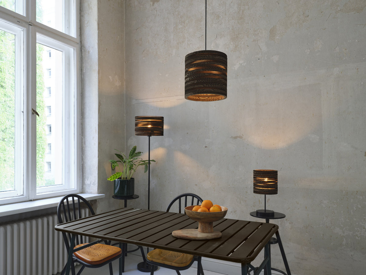 Suspension cylindrique en carton éclairant une salle à manger avec table, chaises et lampadaire.