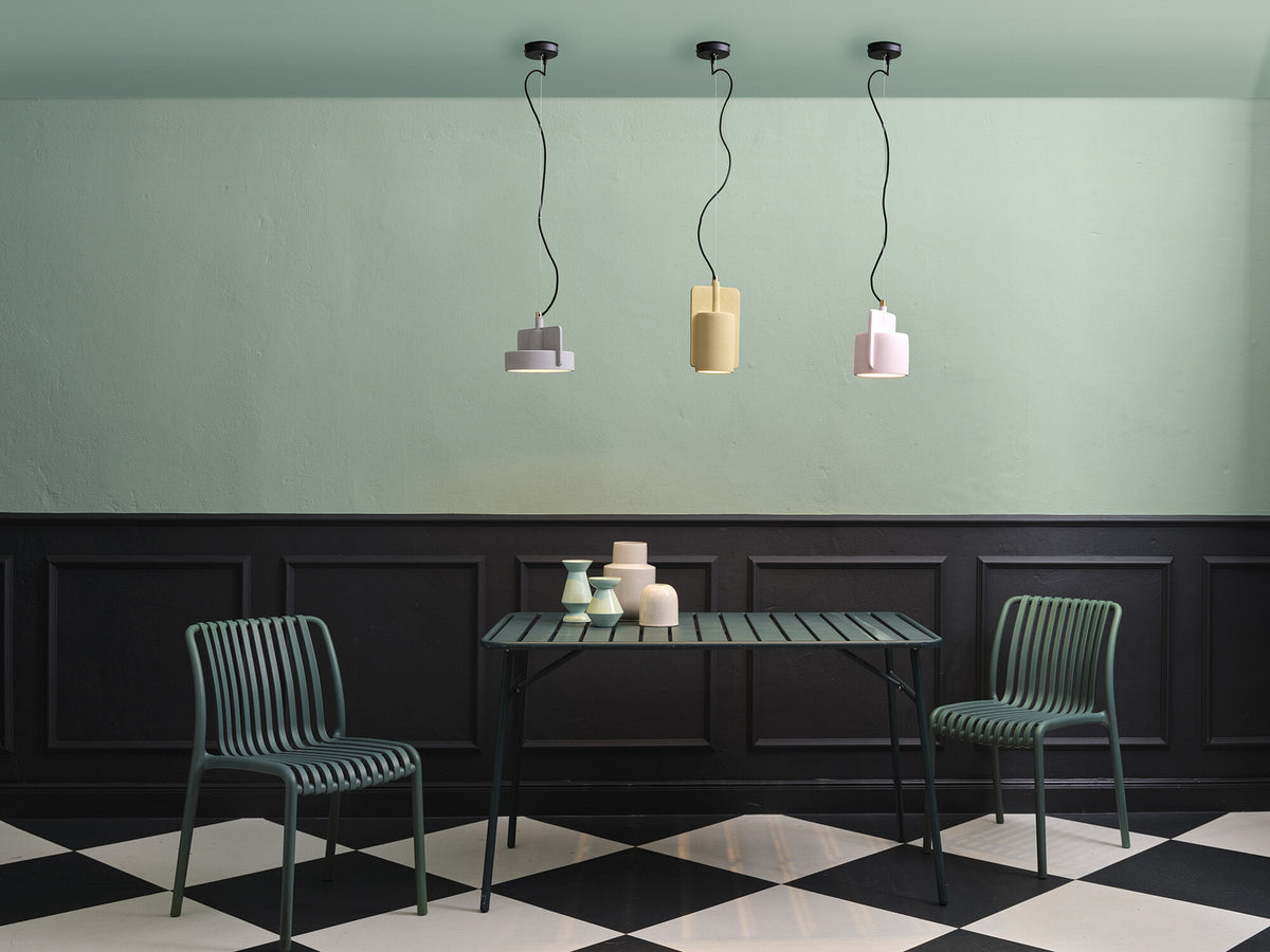 Trois suspensions colorées alignées au-dessus d’une table, chaises vertes et sol en damier.