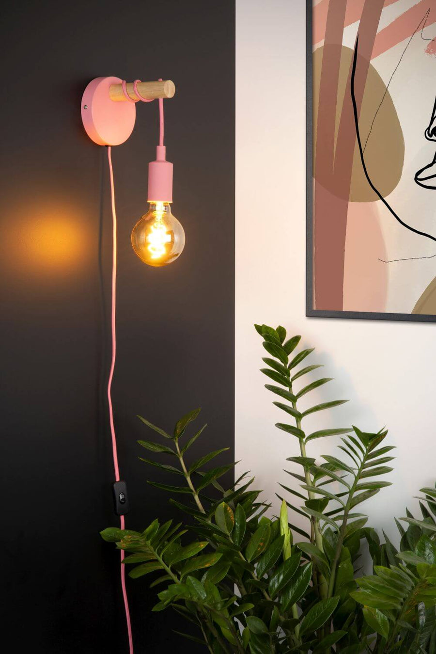 Gros plan sur l'applique murale rose allumée, ampoule visible et plante verte.