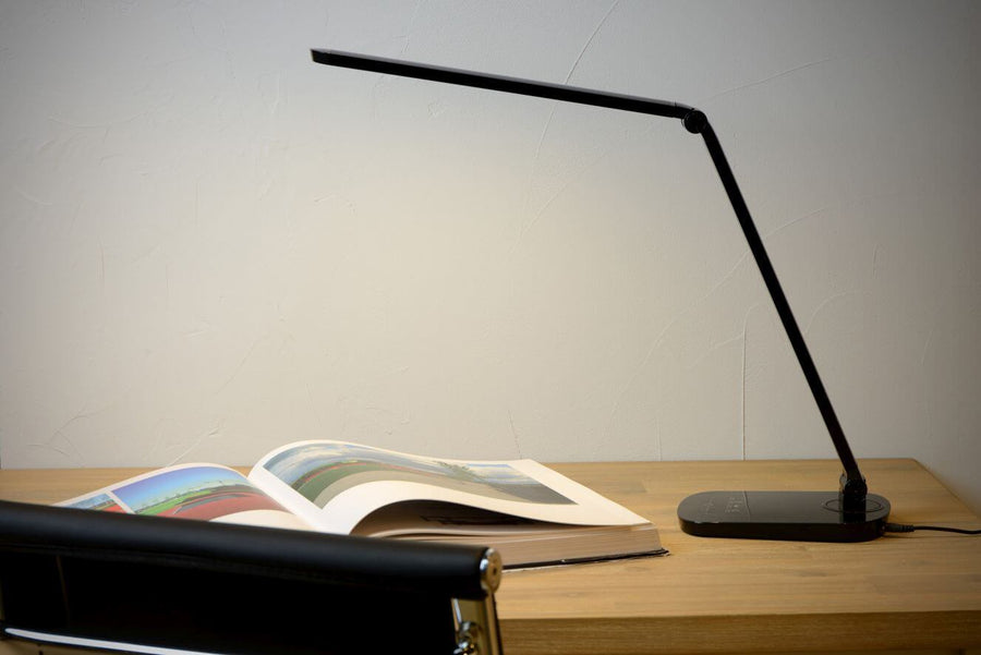 Vue rapprochée de la lampe noire éclairant un magazine ouvert sur une table en bois clair.