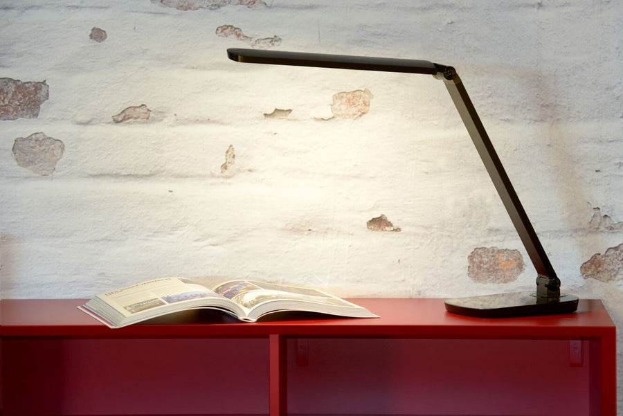 Lampe noire posée sur un meuble rouge, éclairant un livre ouvert devant un mur en briques.