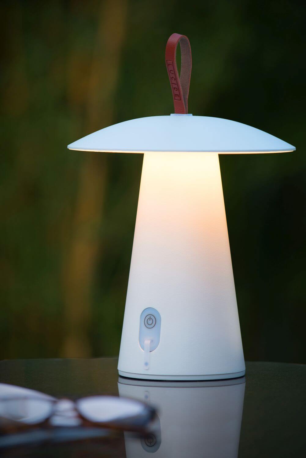 Mise en ambiance nocturne, lampe blanche allumée sur table avec lunettes floues au premier plan.