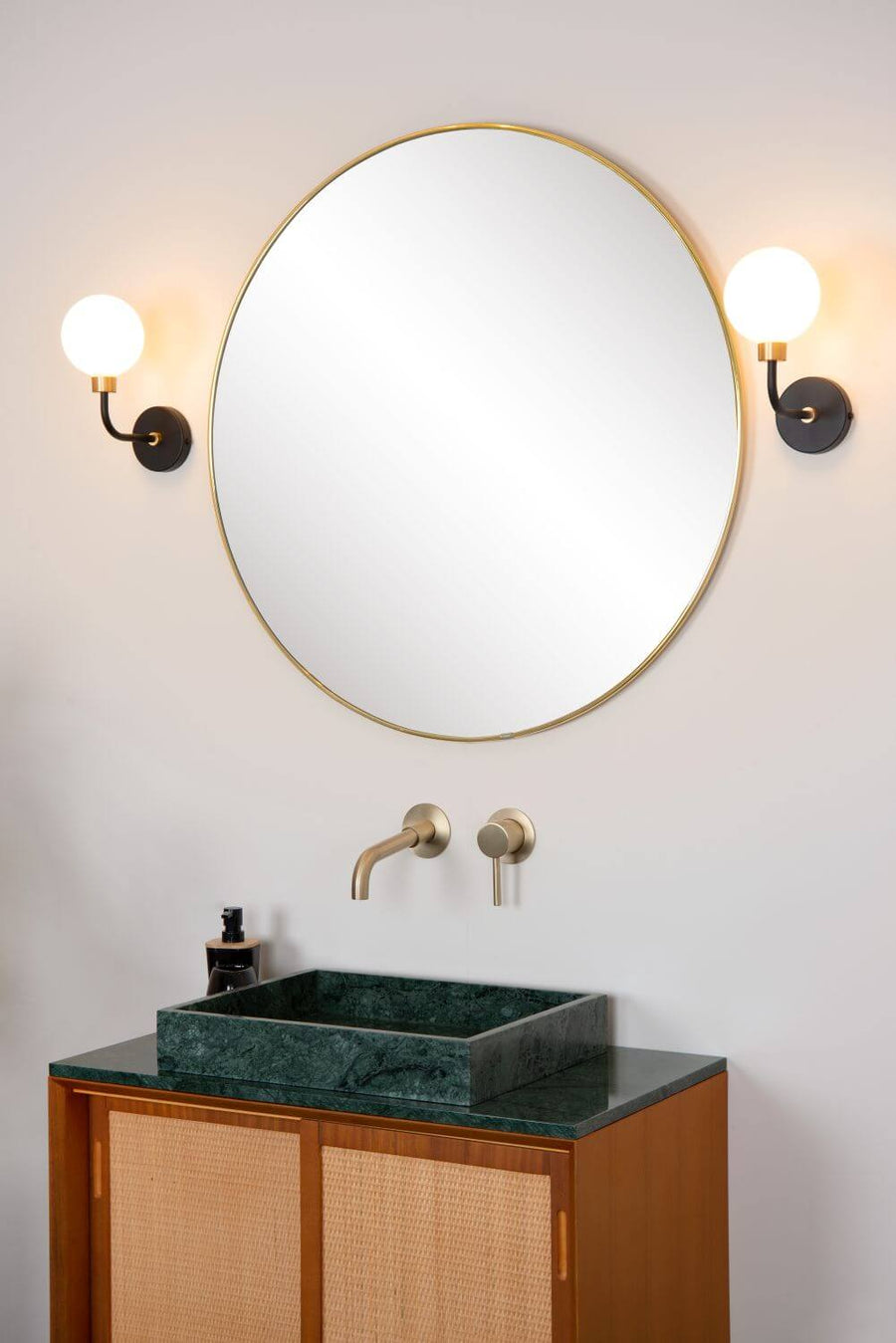 Plan serré sur le miroir rond et la robinetterie dorée, appliques murales non allumées.