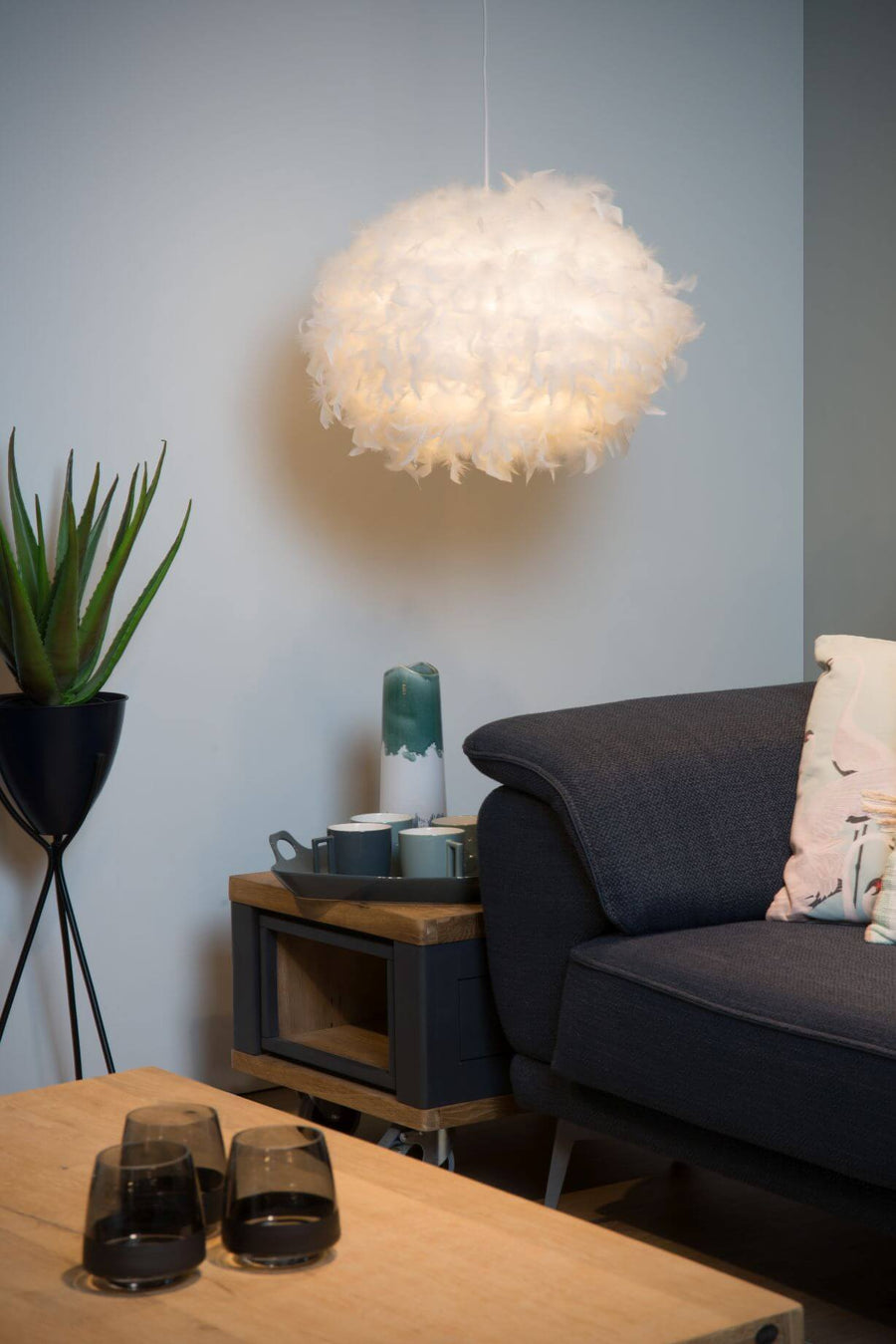Suspension en plumes allumée au-dessus d'un coin salon avec canapé gris et plante verte.