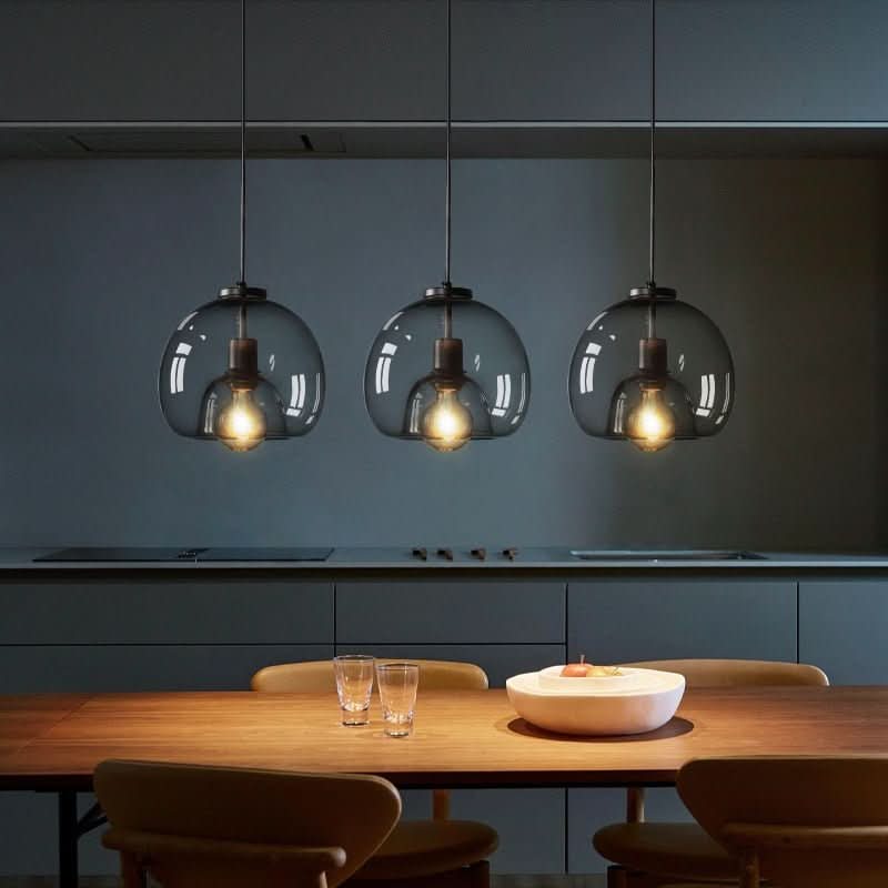 Trois suspensions à globes de verre fumé éclairant une table en bois, cuisine sombre en fond