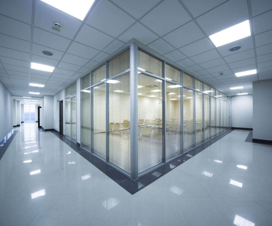 Long couloir de bureaux vitrés avec plafond technique et plusieurs luminaires encastrés allumés.