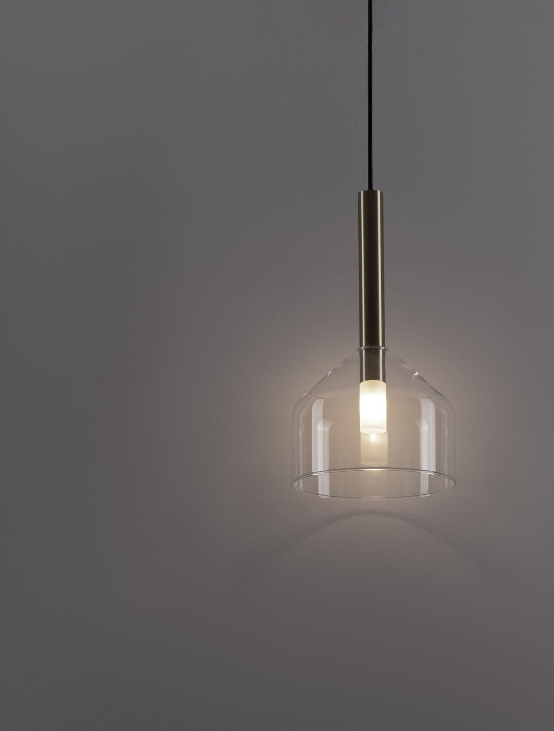 Gros plan sur la suspension allumée, verre transparent et tige dorée, lumière chaude.