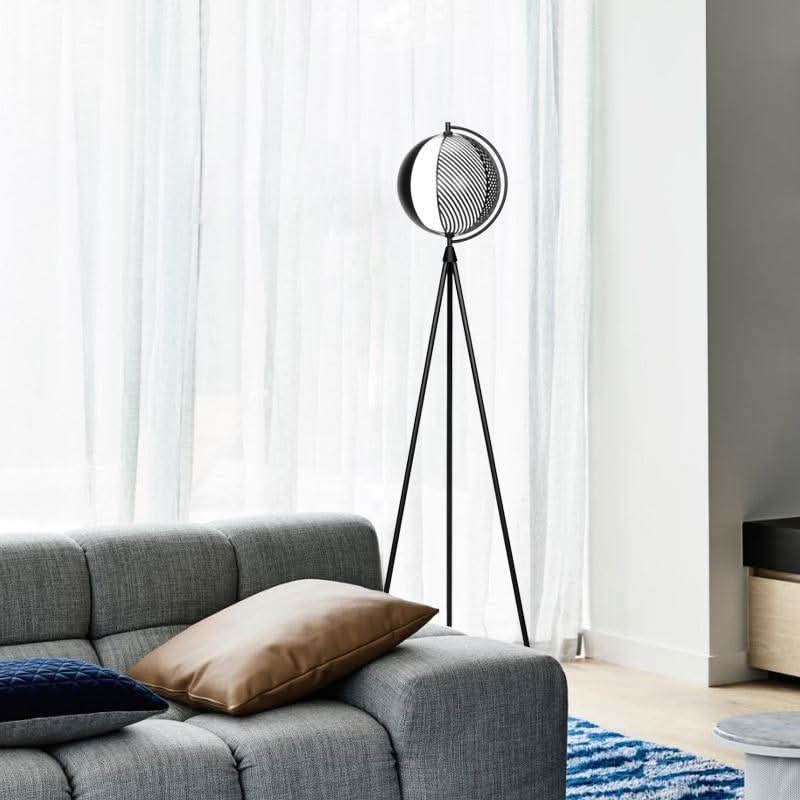 Lampadaire trépied devant rideaux blancs, à côté d'un canapé gris avec coussins