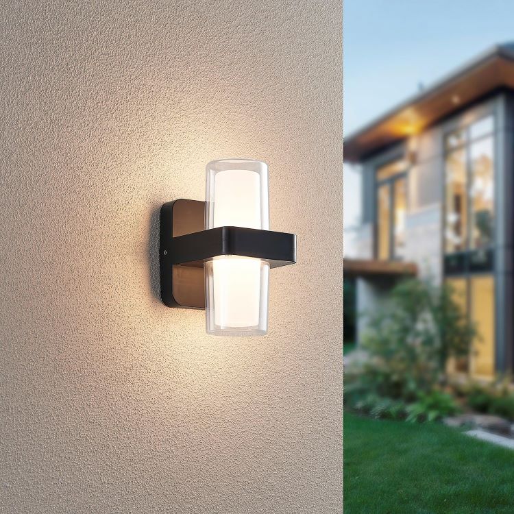 Mise en ambiance extérieure avec applique murale noire allumée sur mur beige et maison floue