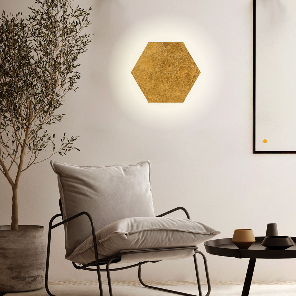 Applique murale hexagonale dorée seule sur un mur clair, halo lumineux diffus au-dessus d'un fauteuil.