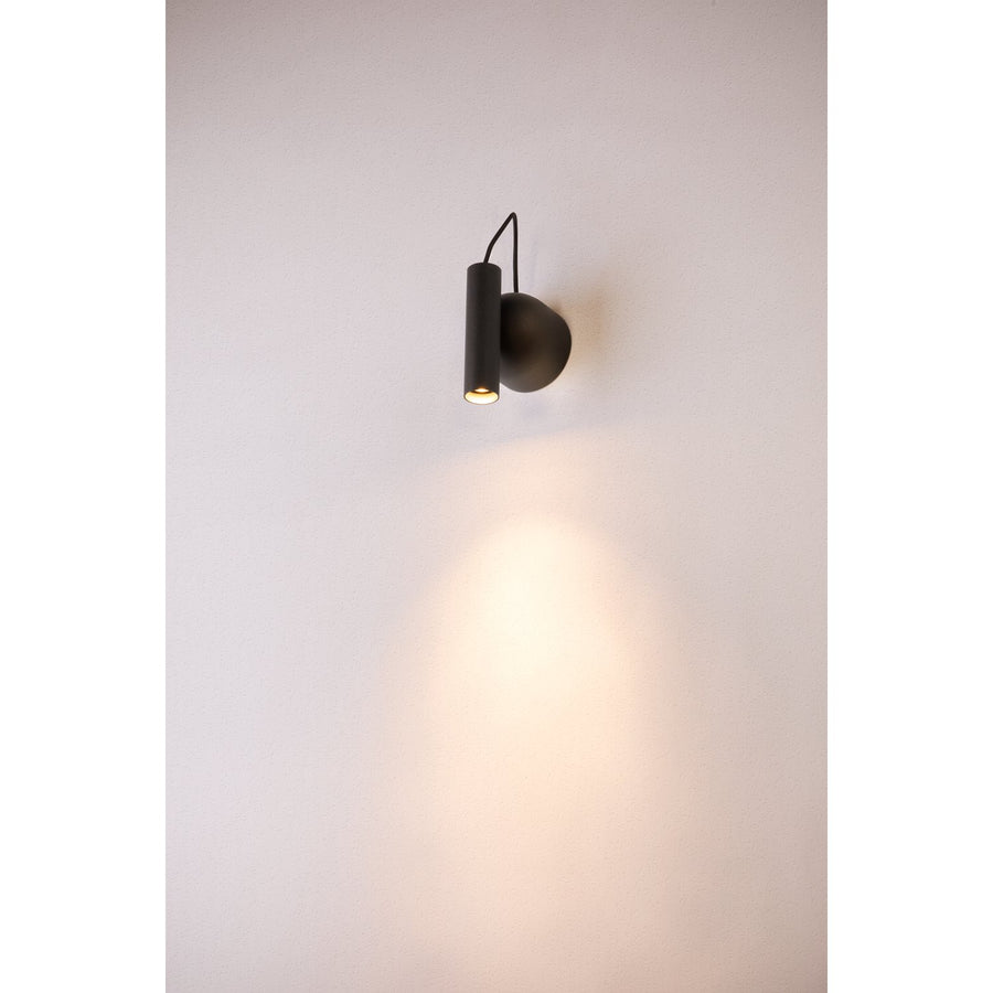 Applique murale noire allumée projetant une lumière chaude et dirigée sur le mur blanc.