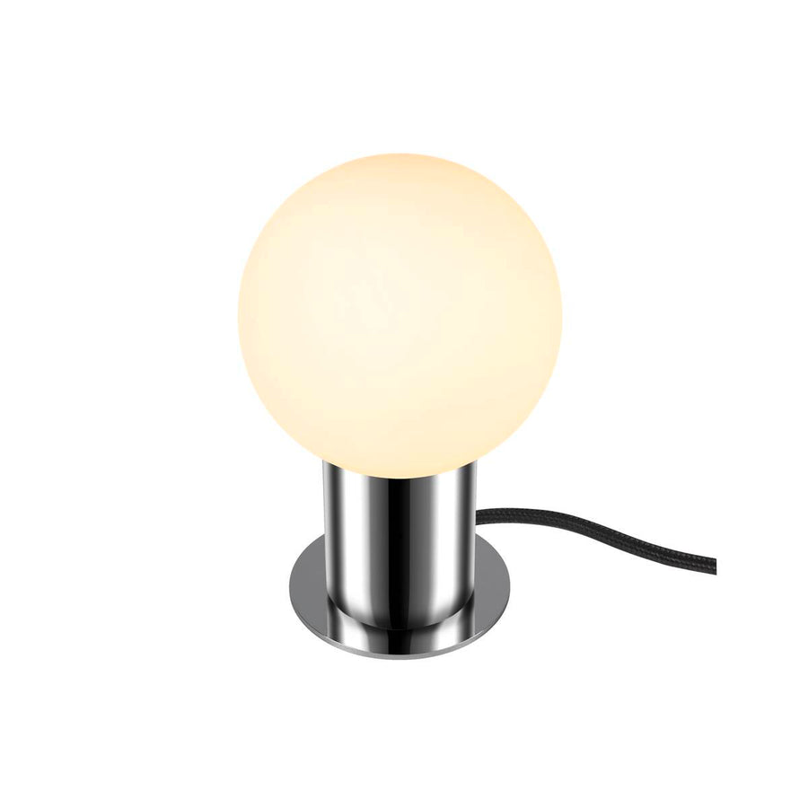 Lampe à poser allumée, globe opalin et base chromée cylindrique sur fond blanc.