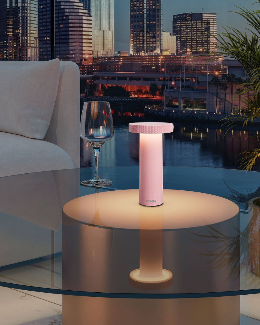 Lampe rose allumée posée sur table basse en verre, vue nocturne urbaine en arrière-plan.