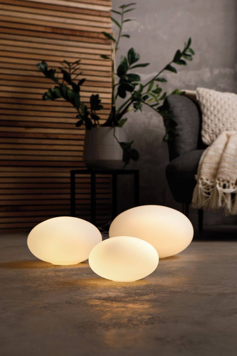 Trois lampes nomades ovales allumées posées au sol près d'un fauteuil, ambiance chaleureuse